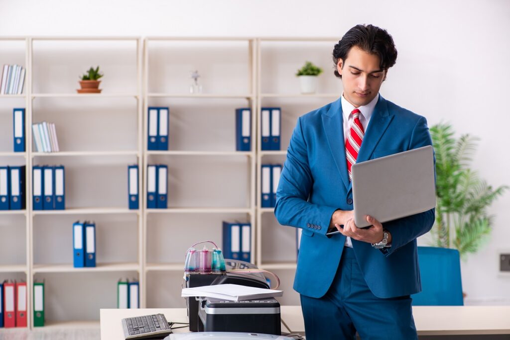 Man in a suit using his laptop next to a copy machine 