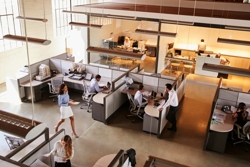A high-angled view of a busy office building. Workers sit in cubicles on their computers while others walk through the office holding papers and folders.