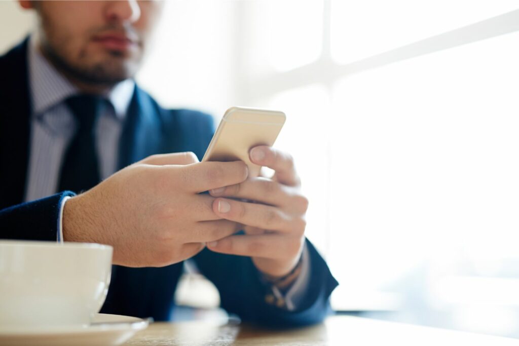 A businessman in a navy blue suit and matching tie sits at his desk with a cup of coffee, sending a fax using his cellphone.