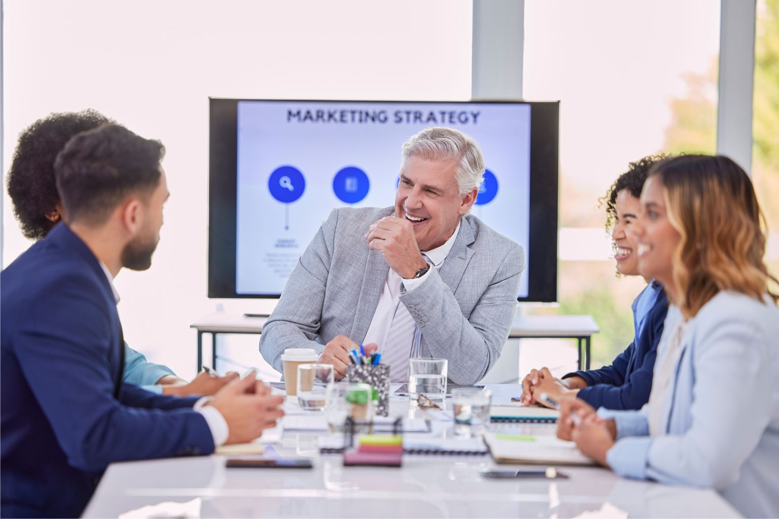image3 A group of five business professionals gathers together around a table to discuss marketing strategy. A screen in the background reads "Marketing Strategy" with blue icons.